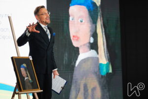 Prowadzący aukcję na scenie pokazuje na licytowany obraz. 