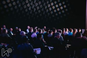 Grupa osób podczas konferencji, ogląda wystąpienie w ciemnej sali. 